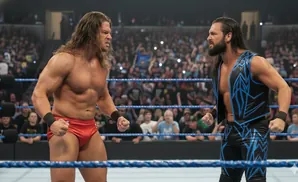 Two wrestlers, one in red trunks and the other in a black and blue outfit, face off in a wrestling ring with a crowd watching.