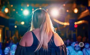 Back view of a woman with blonde hair on stage, facing bright lights and an audience.