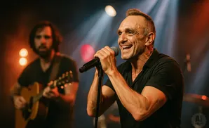 A man sings into a microphone on stage, with another musician playing guitar in the blurred background.