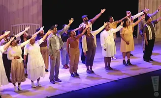 A diverse group of performers stand on stage with arms raised in unison, bathed in stage lighting.