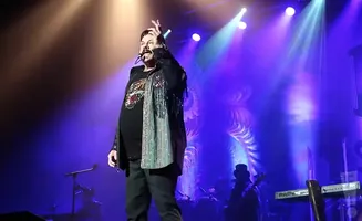 Singer on stage in a black jacket with sparkly fringe and a patterned shirt, performing under purple lights.