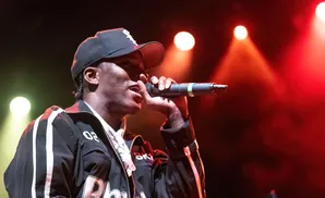 A rapper in a black cap and jacket performs into a microphone under stage lights.