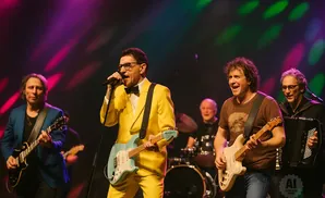 A band performs on stage, with the lead singer in a yellow suit and the guitarist in a brown shirt.