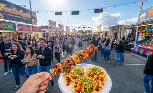 A person holds up a skewer of grilled food and two tacos at a crowded food festival.