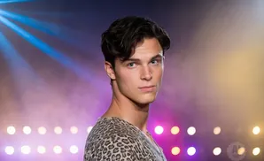 A young man with dark hair and blue eyes looks toward the camera, wearing a leopard-print shirt with a blurred, colorful background.