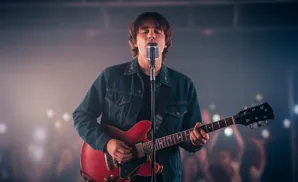 A man in a denim jacket sings into a microphone while playing a red electric guitar on stage.
