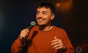 A man with short brown hair smiles while holding a microphone and wearing an orange sweatshirt.