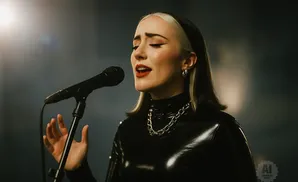 A woman with blonde streaks in her hair sings into a microphone while wearing a black latex top and a chunky silver necklace.
