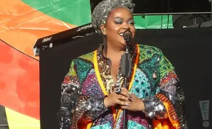 Jill Scott wearing a brightly patterned jacket and silver headwrap sings into a microphone on stage.