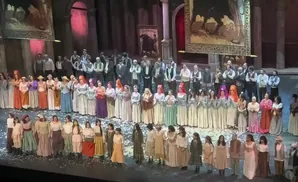 A large cast stands on stage, a mix of men and women in period costumes, bowing to the audience amidst scattered confetti.