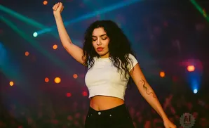 Singer on stage with fist raised, wearing a white crop top and black pants, with colorful stage lights in the background.