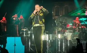 A male singer in a decorated charro suit performs on stage with a mariachi band.