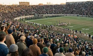 Spectators in vintage clothing watch a football game from a stadium, with players on the field and a marching band.