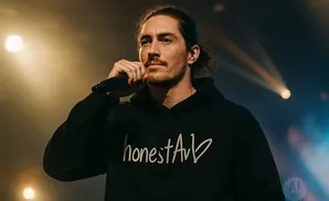 A man with long brown hair tied back, wearing a black hoodie, holds a microphone to his mouth on a stage lit by warm spotlights.