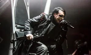 Marilyn Manson sings into a microphone on stage, dressed in black leather and makeup.