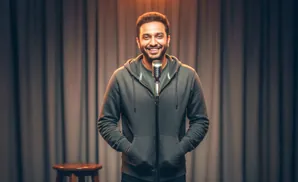 A smiling man in a grey hoodie stands at a microphone on a stage with curtains.