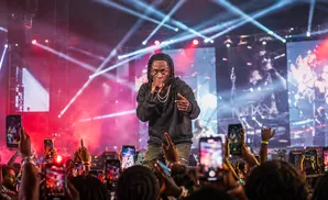 A rapper performs on stage with bright lights and a crowd holding up phones.
