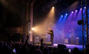 A comedian on stage with blue and orange lights, speaking to an audience.