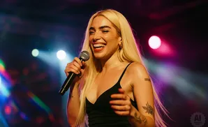 A woman with long blonde hair sings into a microphone on stage, illuminated by colorful stage lights.