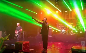 A bald man plays guitar on stage while a singer points and sings under bright green and yellow lights.