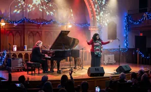 A man plays a grand piano on a stage decorated for Christmas, with an audience in the foreground.