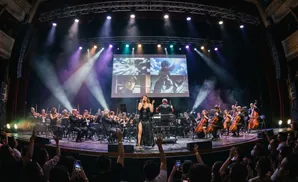 Singer and orchestra perform in front of a screen displaying anime clips.