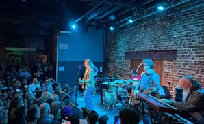 Musicians perform on a dimly lit stage with a brick wall background, in front of a cheering crowd.