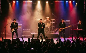 A band performs on stage with red and blue lights, a drummer behind them, and a cheering crowd in the foreground.