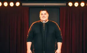 A man in a black shirt stands before a microphone on a stage with red curtains and bright spotlights.
