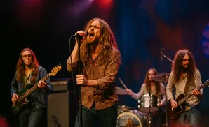 A male singer with long, wavy brown hair sings into a microphone on stage, flanked by bandmates playing guitar and drums.