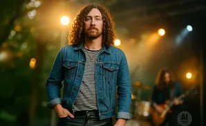 A man with curly brown hair and a beard in a denim jacket and striped shirt stands with his hand in his pocket.