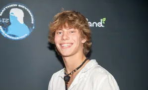 A young man with light brown hair and blue eyes smiles at the camera, wearing a white shirt and a beaded necklace with a pendant.