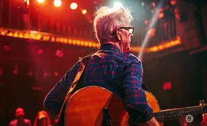 A man with white hair and glasses plays an acoustic guitar on a stage lit by red lights.