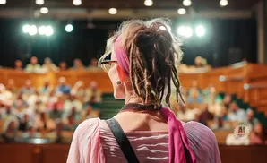Woman with dreadlocks and a pink headscarf facing away from the camera, speaking to an audience in an auditorium.