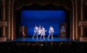 A ballet performance on a blue stage with five dancers in white costumes, with an orchestra and audience in the foreground.