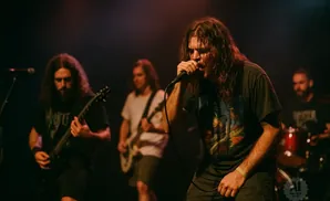 A rock band performs live, with the lead singer in the foreground singing into a microphone.