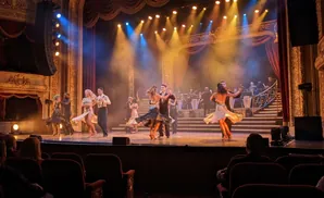 Dancers perform on a smoke-filled stage in a grand theatre with elaborate gold decorations and red curtains.