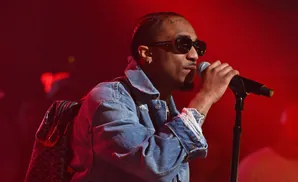A man with sunglasses and a denim jacket sings into a microphone on a red-lit stage.