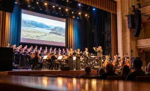 A choir and orchestra perform on stage in front of a large screen displaying a landscape.