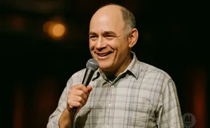 A bald man in a plaid shirt holds a microphone and smiles.