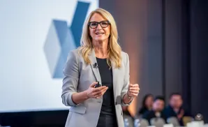 Woman in a grey suit and glasses speaks at a conference, holding a clicker.