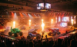 Monster trucks with flames erupting around them perform in a stadium with a large crowd.