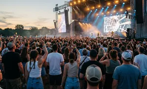 A large crowd cheers at a concert during sunset, with musicians performing on a stage with bright lights.