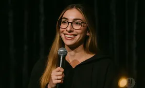 Woman with glasses and long blonde hair smiling and holding a microphone.