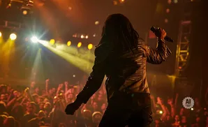 Singer with dreadlocks in a leather jacket performs for a crowd at a concert, illuminated by stage lights.