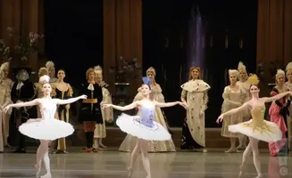 Ballet dancers in colorful tutus on stage with an ensemble of costumed performers.