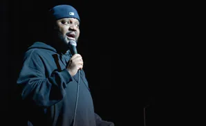 A man in a black hoodie and baseball cap performs on stage with a microphone in hand.