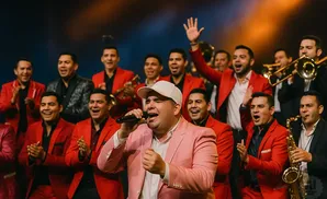 A male singer in a pink suit sings into a microphone, with a band in red suits behind him playing instruments.