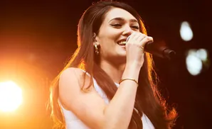 A woman with long brown hair smiles while singing into a microphone on a stage.