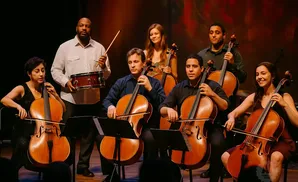 An orchestra of cellists and a drummer perform on stage.